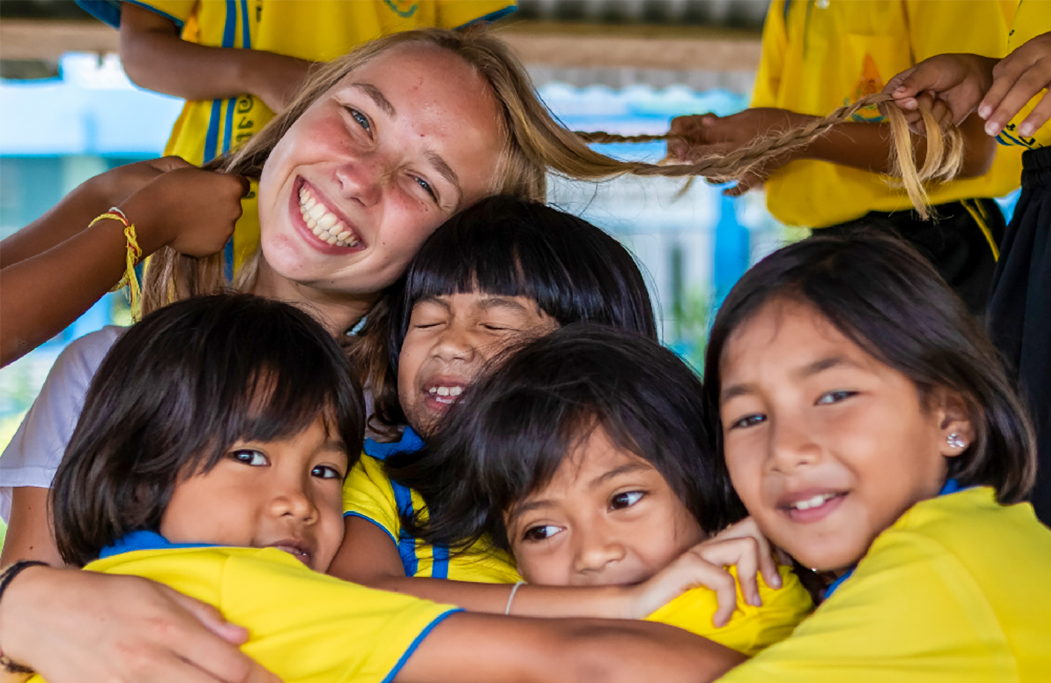 A joyful volunteer embraces smiling kindergarten children who playfully braid her hair in Thailand.