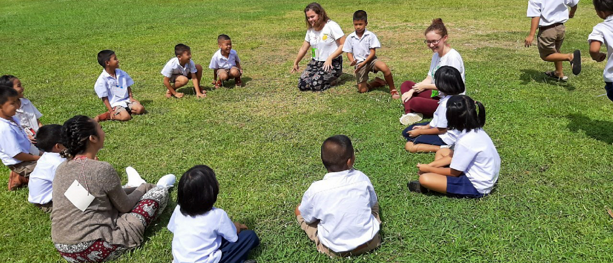 Two volunteers teach a group of young Thai children sitting in a circle on green grass.
