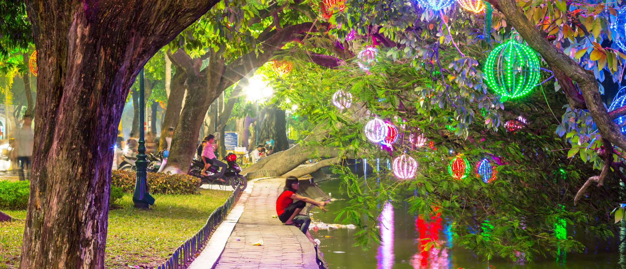 A person sits by water under trees adorned with colorful glowing lanterns in Hanoi.