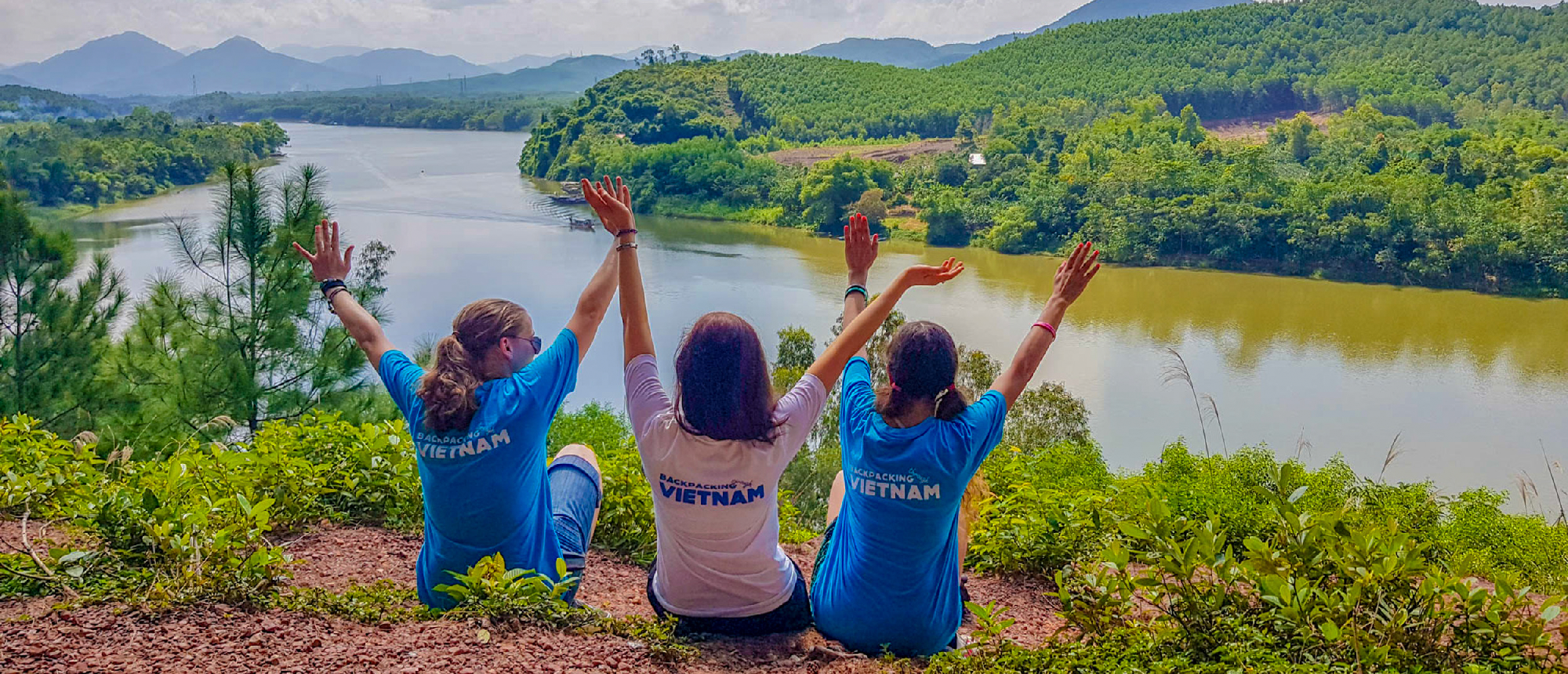 Three women with 'Vietnam' shirts joyfully raise arms overlooking the Perfume River near Hue.