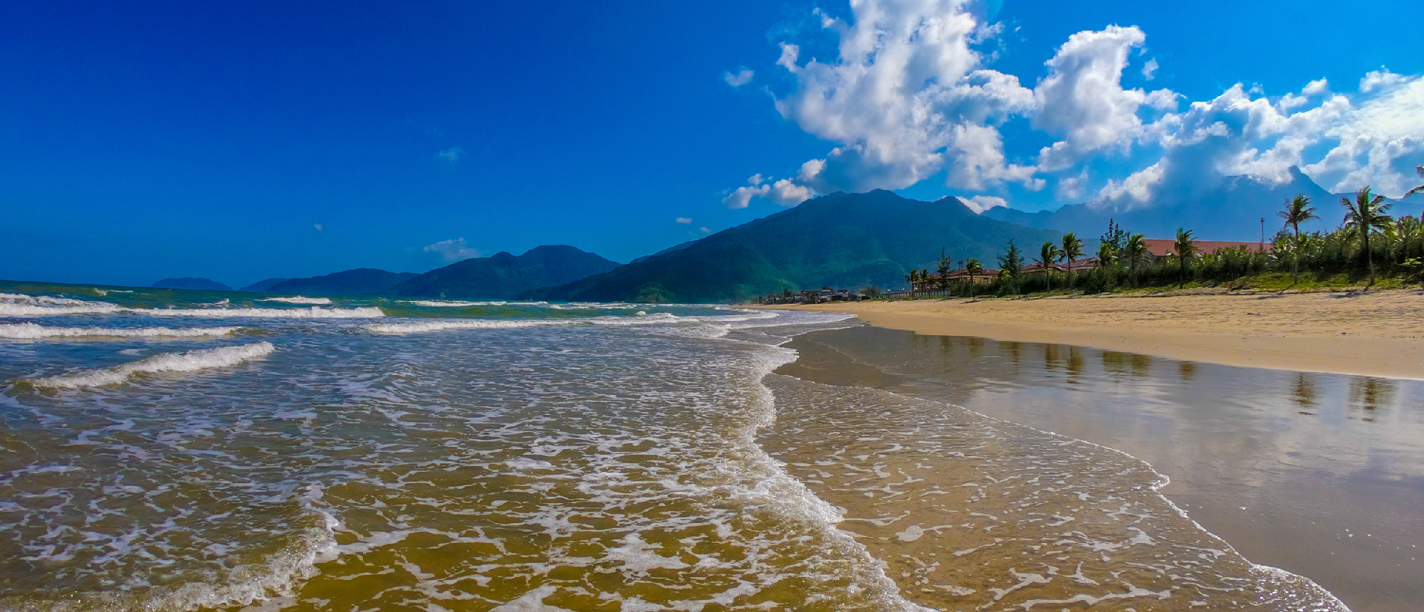 Gentle waves wash onto the golden sands of Nha Trang beach, backed by mountains and palm trees.
