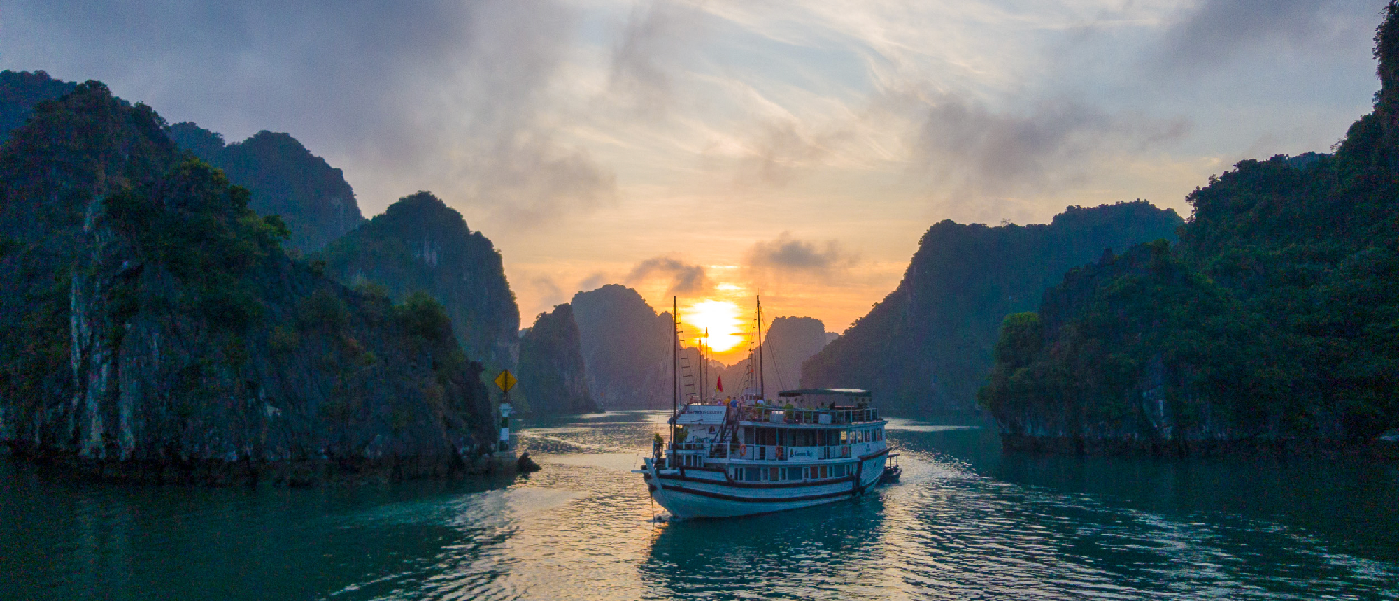 A tour boat sails through the dramatic karsts of Halong Bay during a golden sunset.