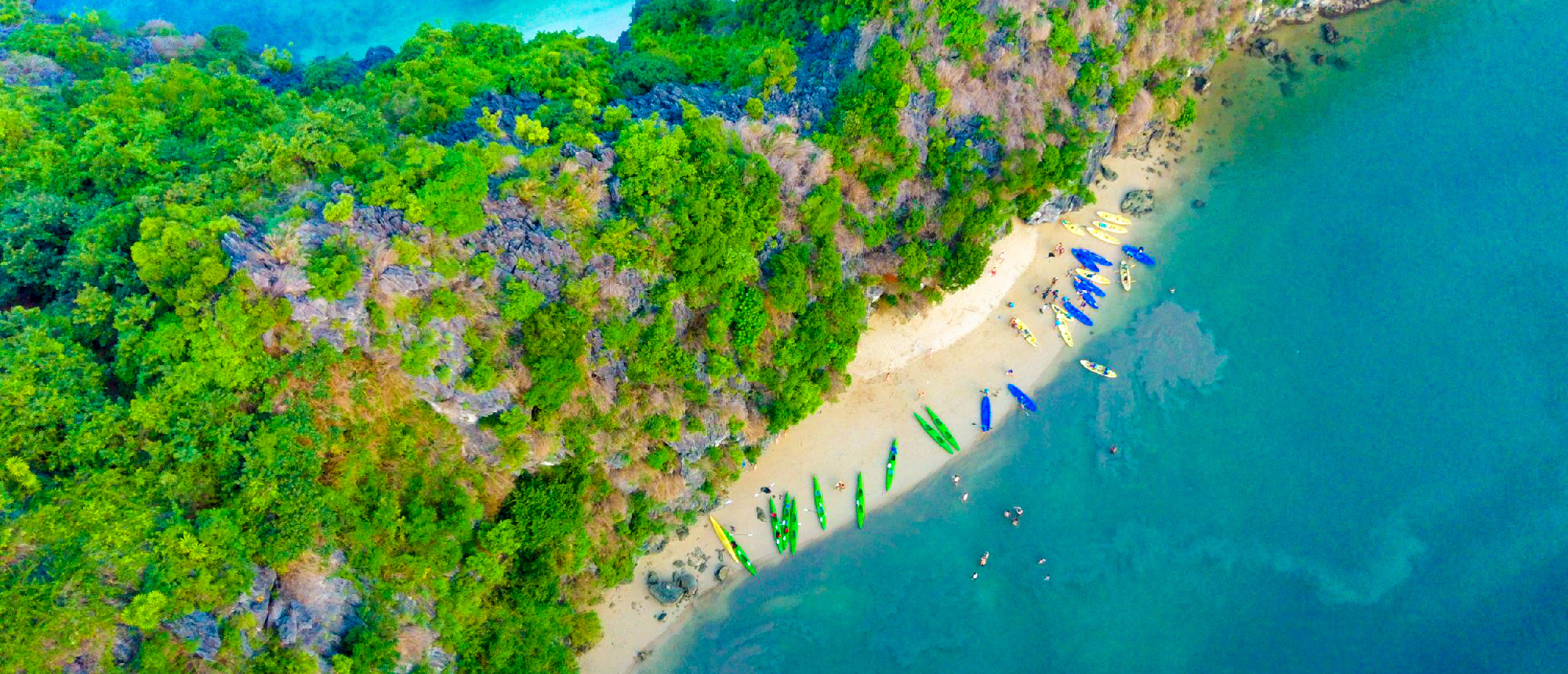 Colorful kayaks rest on a sandy beach beside a vibrant green island in Halong Bay.