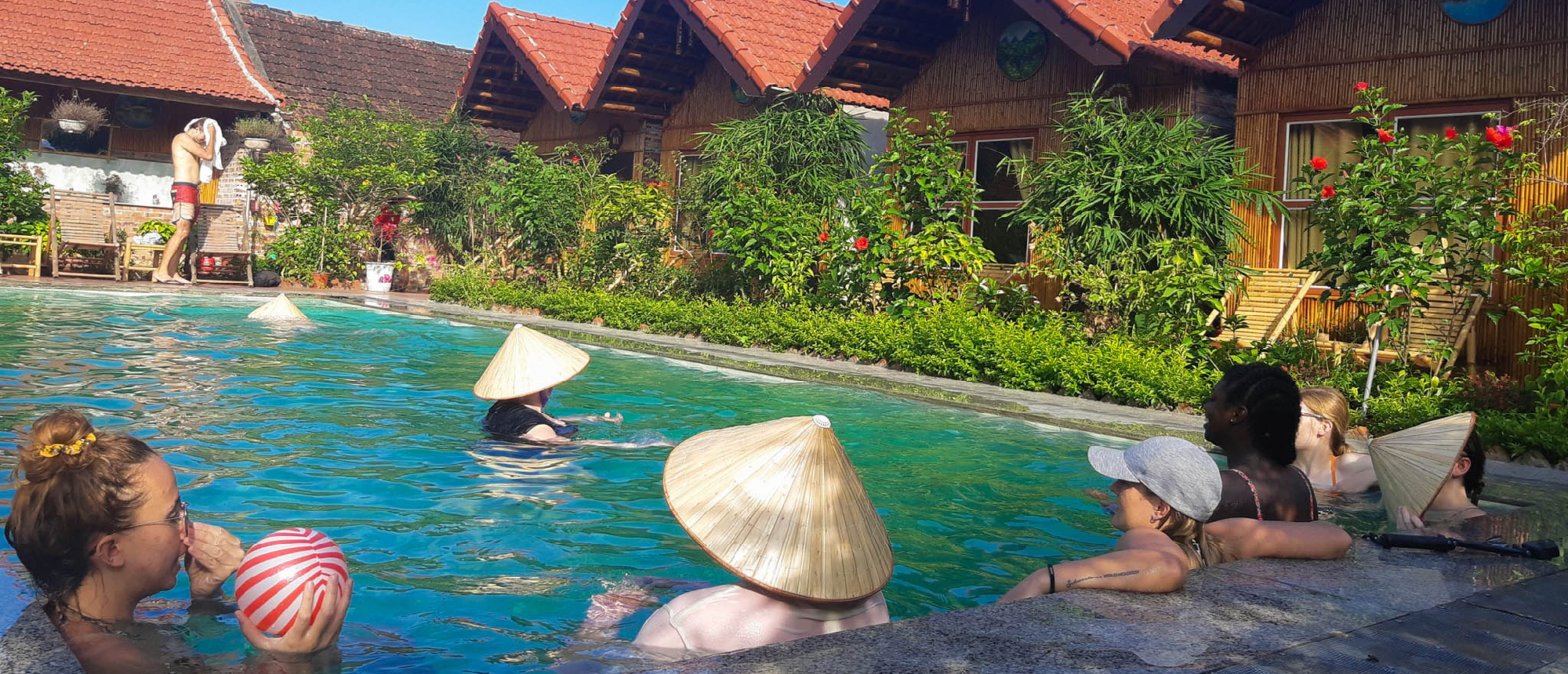 Travelers relax in a swimming pool, some wearing conical hats, with charming bungalows in Vietnam.