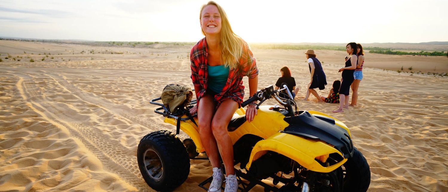 Smiling blonde woman on a yellow ATV with friends on Mui Ne sand dunes, Vietnam.