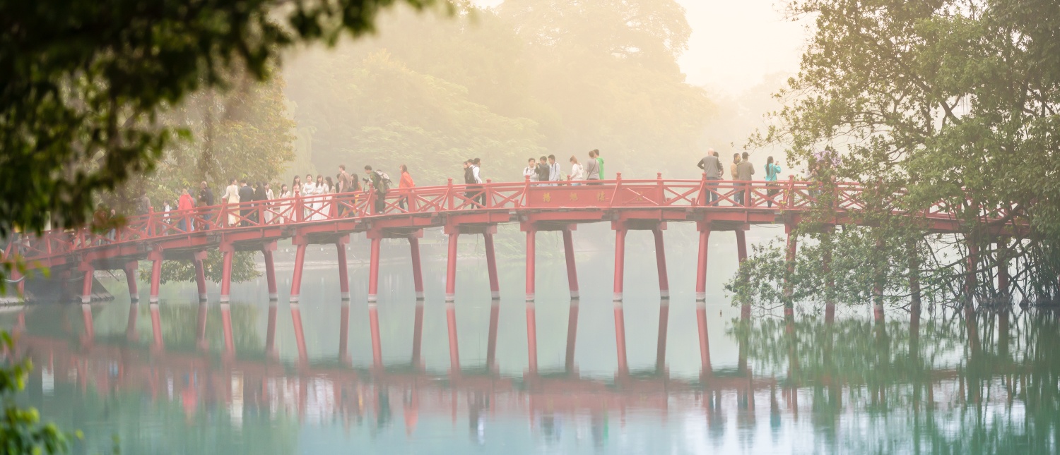 People stroll across the iconic red Huc Bridge over Hoan Kiem Lake in Hanoi, Vietnam, on a misty morning.