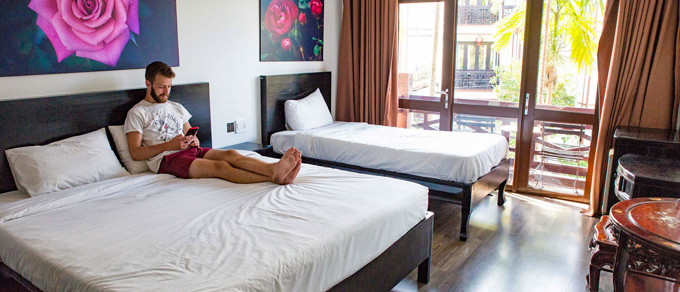 A man with a beard sits on a large bed, scrolling on his phone in a hotel room.