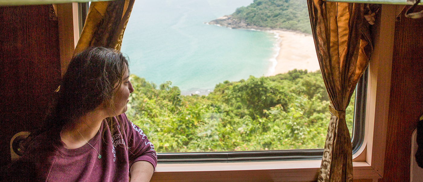 A woman looks out a train window at a beautiful sandy beach and turquoise ocean.