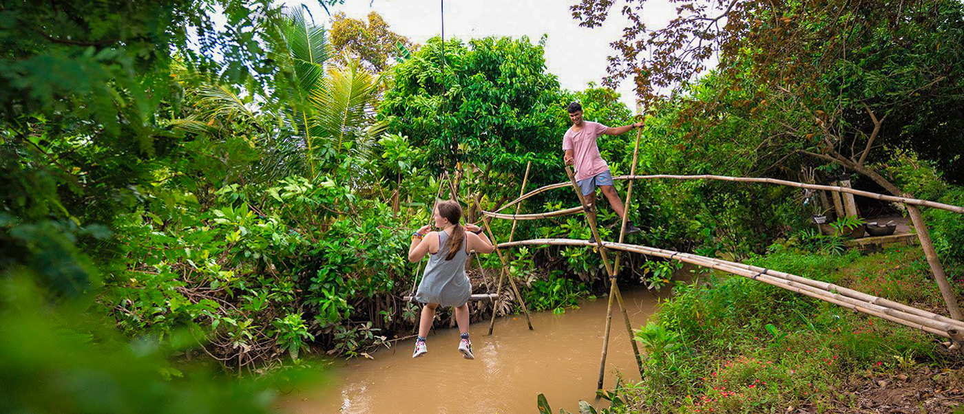 Adventurous travelers balance on a rickety bamboo bridge above a muddy river in the Mekong Delta.