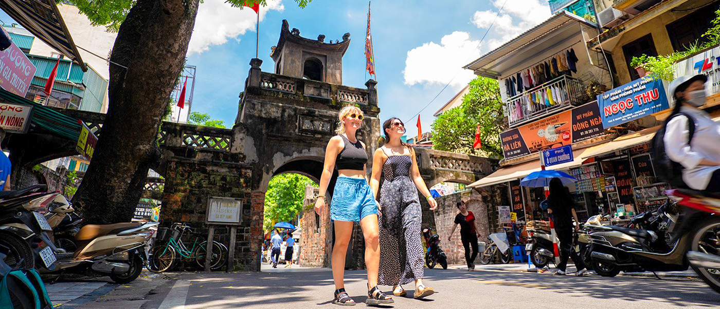Smiling women walk past an ancient gate and busy shops on a sunny day in Hanoi.