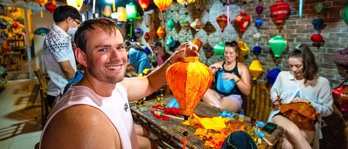 A smiling man holds his handmade orange lantern in a vibrant Hoi An workshop with other travelers.