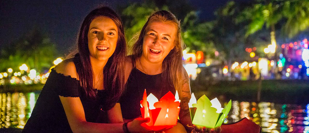 Smiling women holding colorful lanterns by the illuminated river in Hoi An at night.