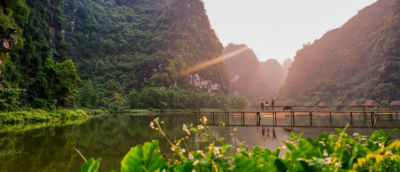 People and a water buffalo cross a wooden bridge over a calm lake at sunset in Ninh Binh.