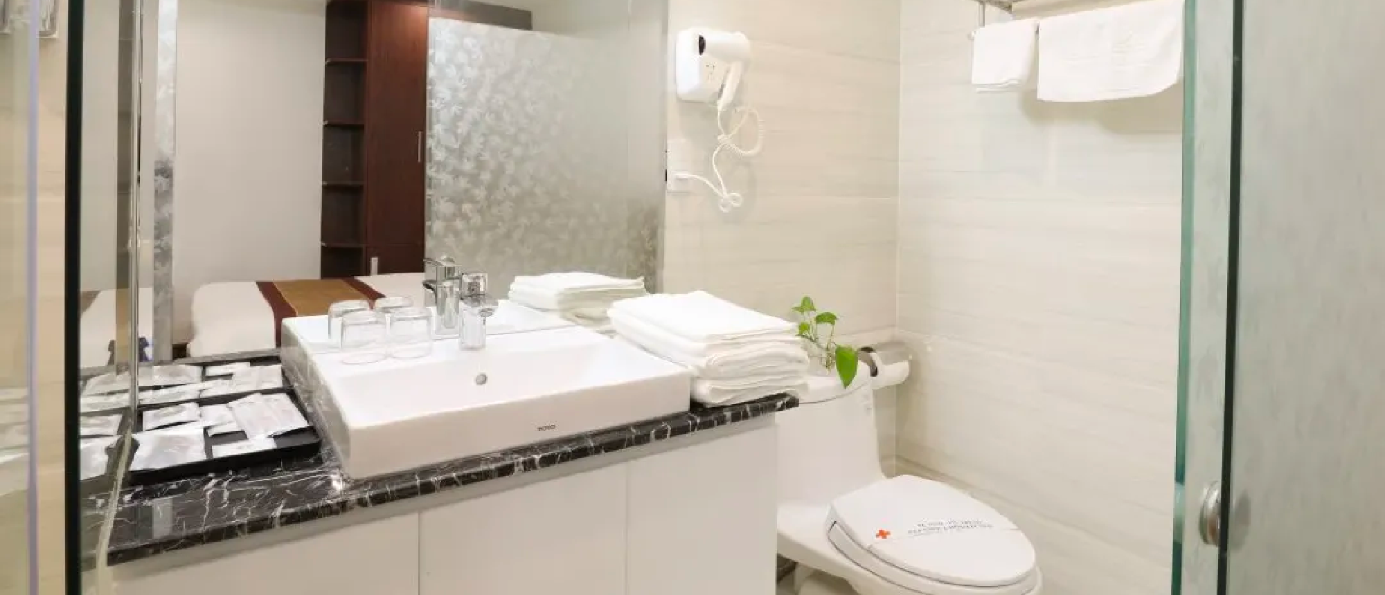 A bright, modern hotel bathroom in Ho Chi Minh with a sink, toilet, fresh towels, reflecting a bed.