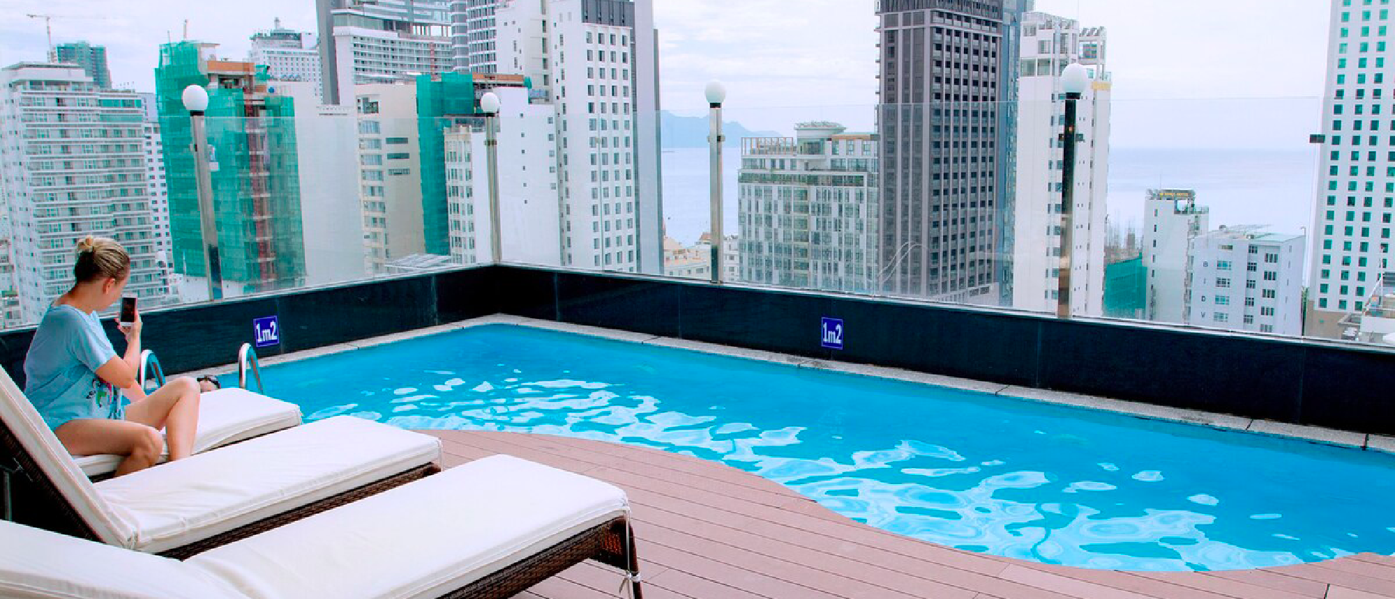 A person sits by a rooftop pool, checking their phone, with Nha Trang city and ocean views.