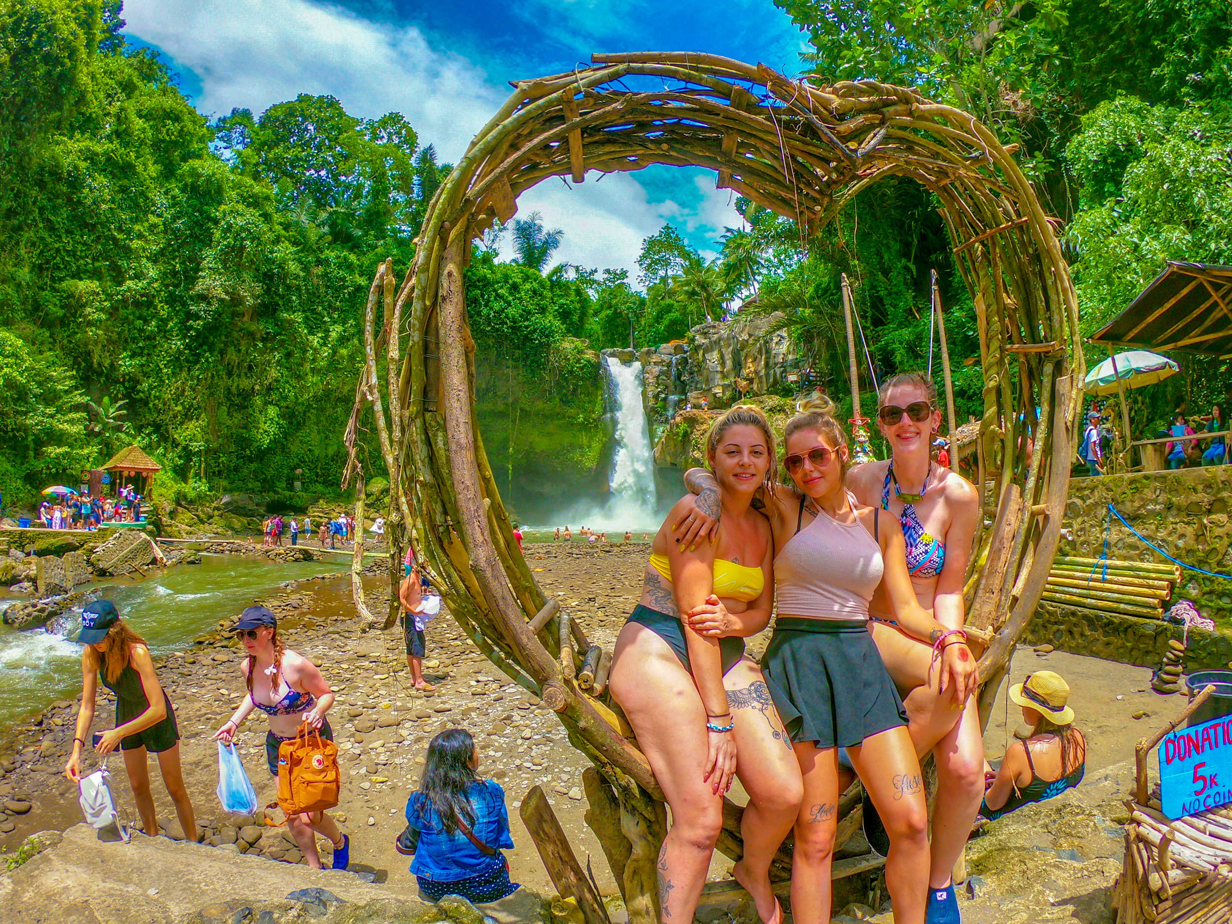 Three women pose in a heart-shaped wooden frame at a beautiful waterfall in Bali.