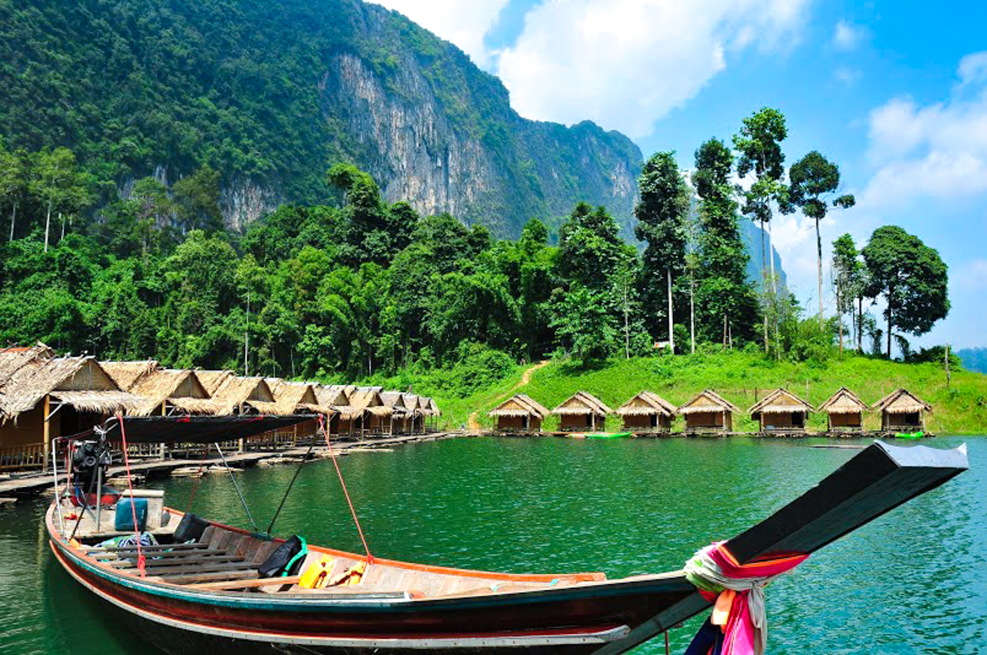 A traditional longtail boat on a green lake, with floating bungalows and lush mountains in Thailand.
